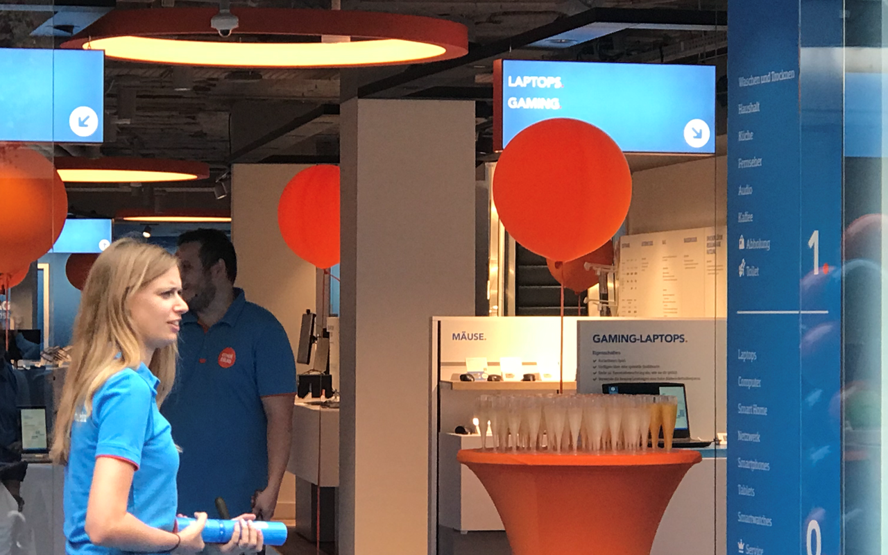Am Eingang des blau gefärbten Geschäftes von Coolblue steht ein orangefarbiger Tisch für den Sektempfang