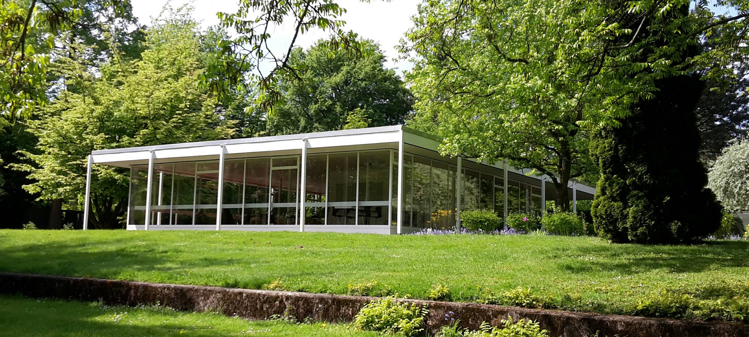 Der weiße gläserne Lesepavillon im Grugapark von Wiese und Bäumen umgeben.