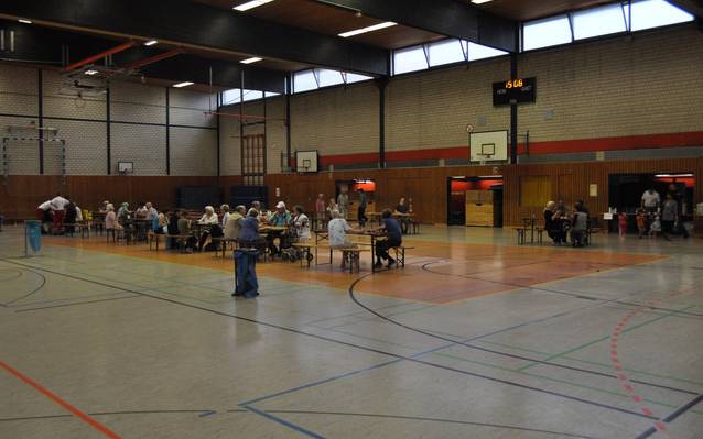 Mehrere Menschen sitzen in der Turnhalle der Schule an der Wolfskuhle in Essen an Tischen und warten.