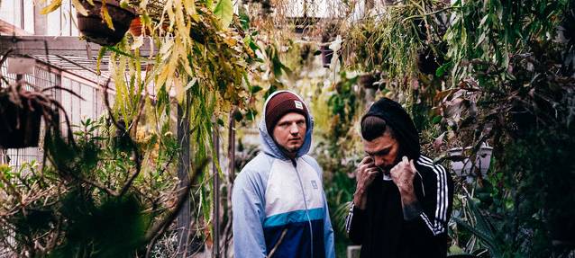 Zwei Jungs in Sportjacken und Mützen stehen zwischen Büschen, sie sind Alidaxo.