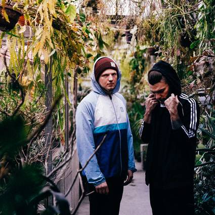 Zwei Jungs in Sportjacken und Mützen stehen zwischen Büschen, sie sind Alidaxo.