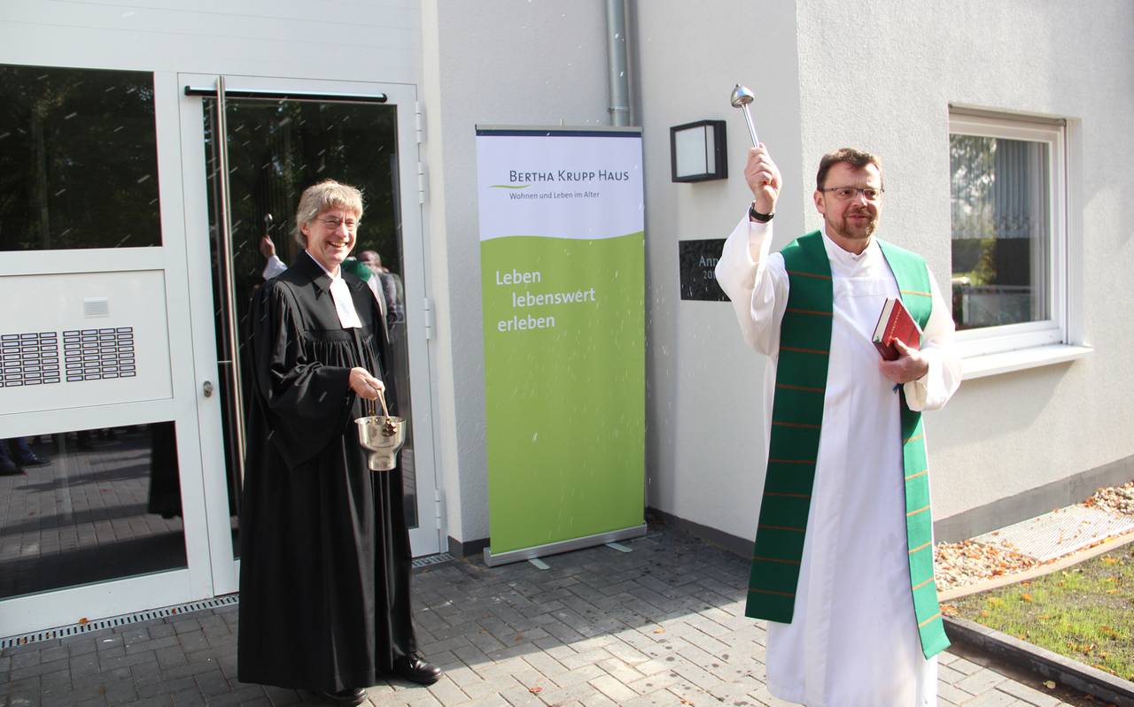 Vor dem Eingang zu einem Altenheim stehen ein zwei Pfarrer, die bei der Eröffnung das Altenheim segnen.