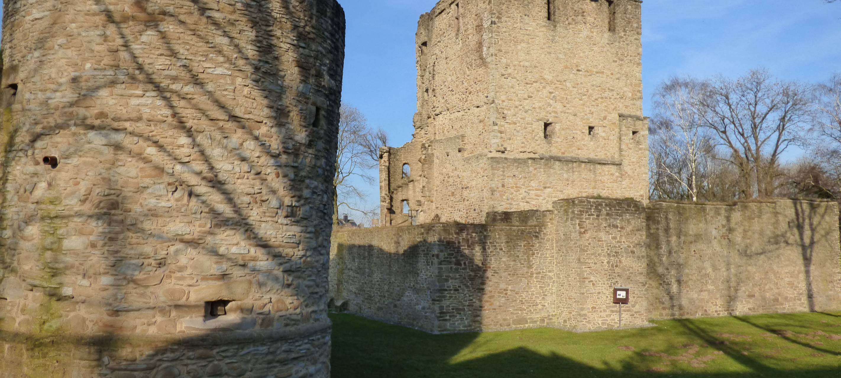 Die graue Burgruine in Burgaltendorf mit starken Mauern, Türmen und Wiesen drum herum.