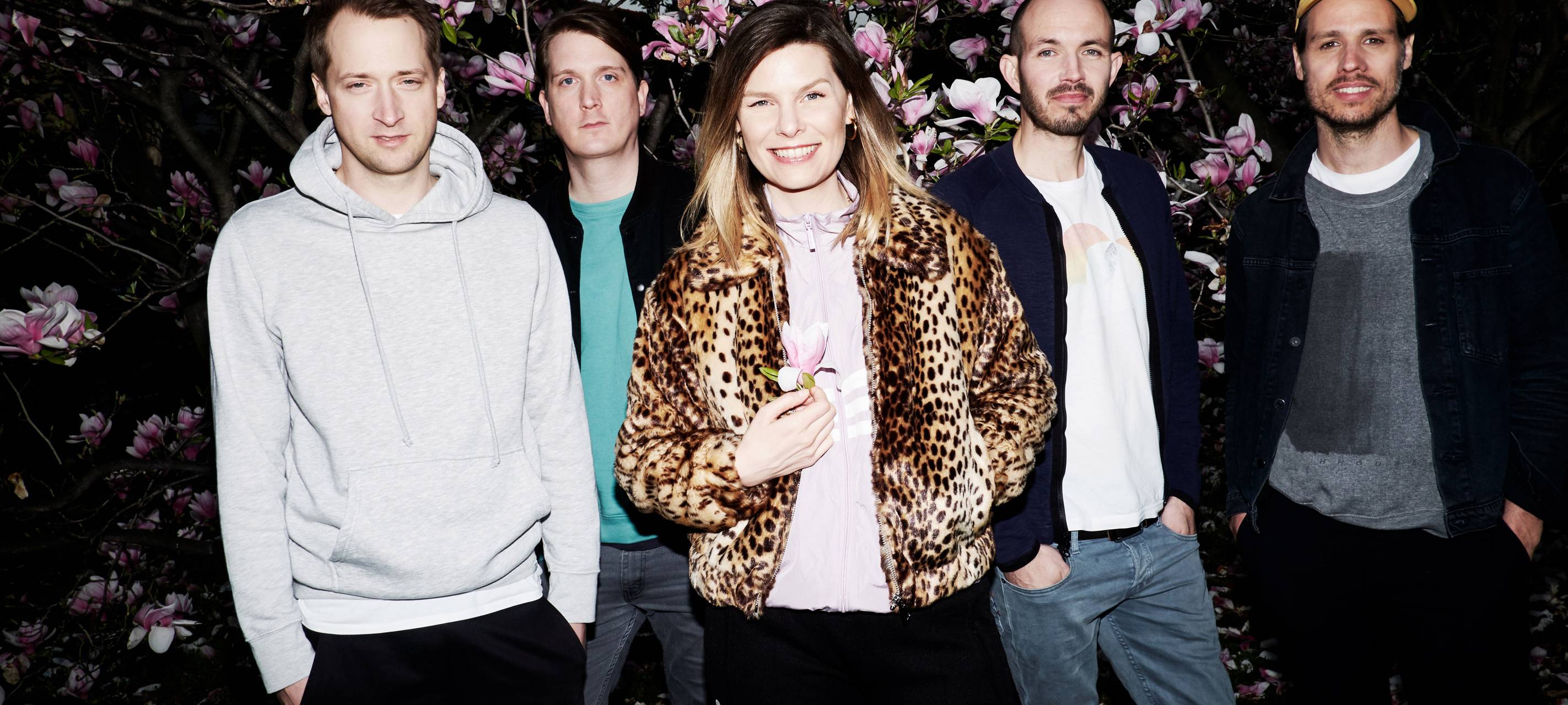 Die Band Juli mit vier Männern und der Frontfrau in der Mitte vor einem Busch mit rosa Blüten.