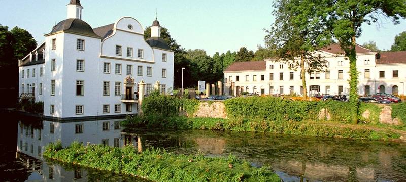 Das Schloss Borbeck ist ein weißes großes Schloss mit hohen Fenstern, direkt an einem kleinen See gelegen mit einem Park dahinter.