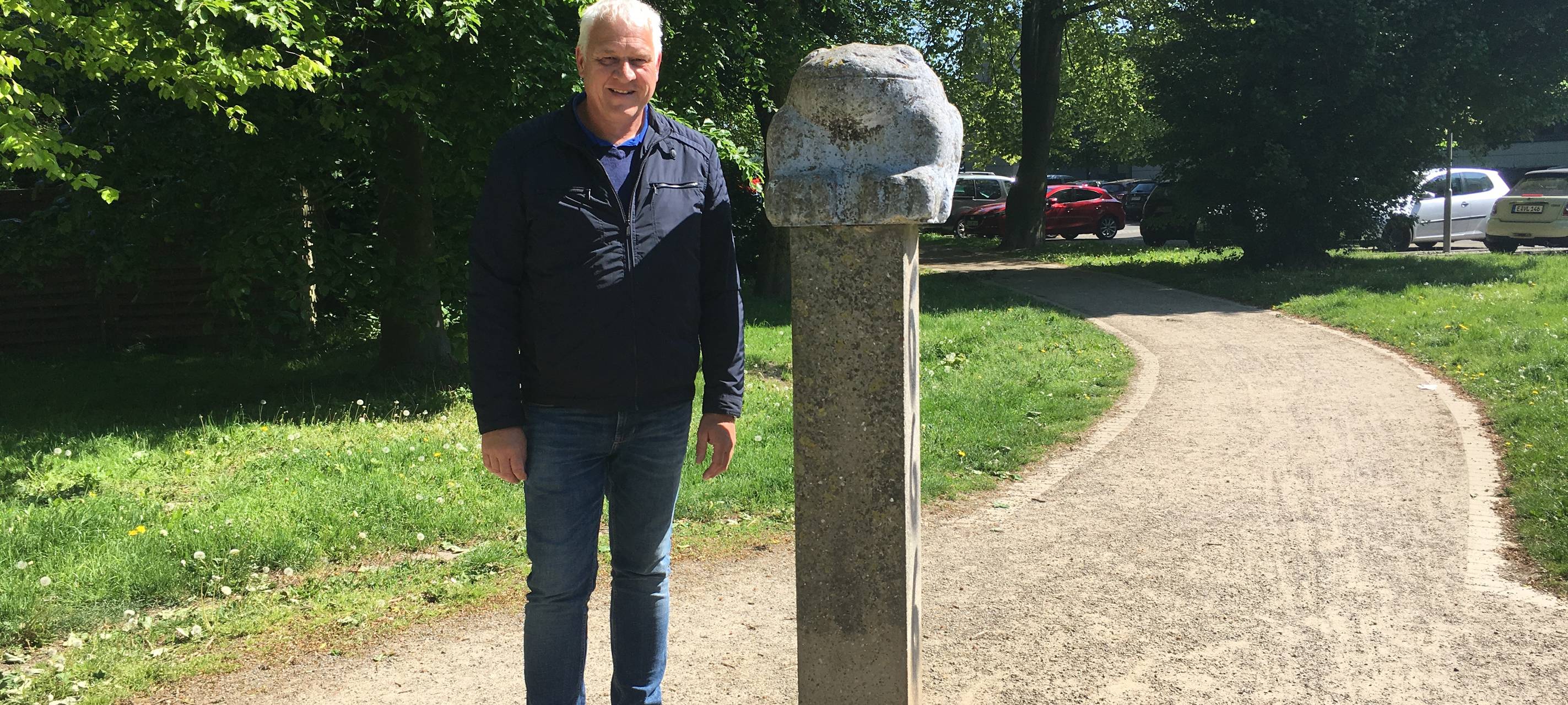 Ein älterer Mann, Landschaftsarchitekt Ulrich Falke, steht neben einem Steinfrosch auf einem Sockel in Schönebeck.