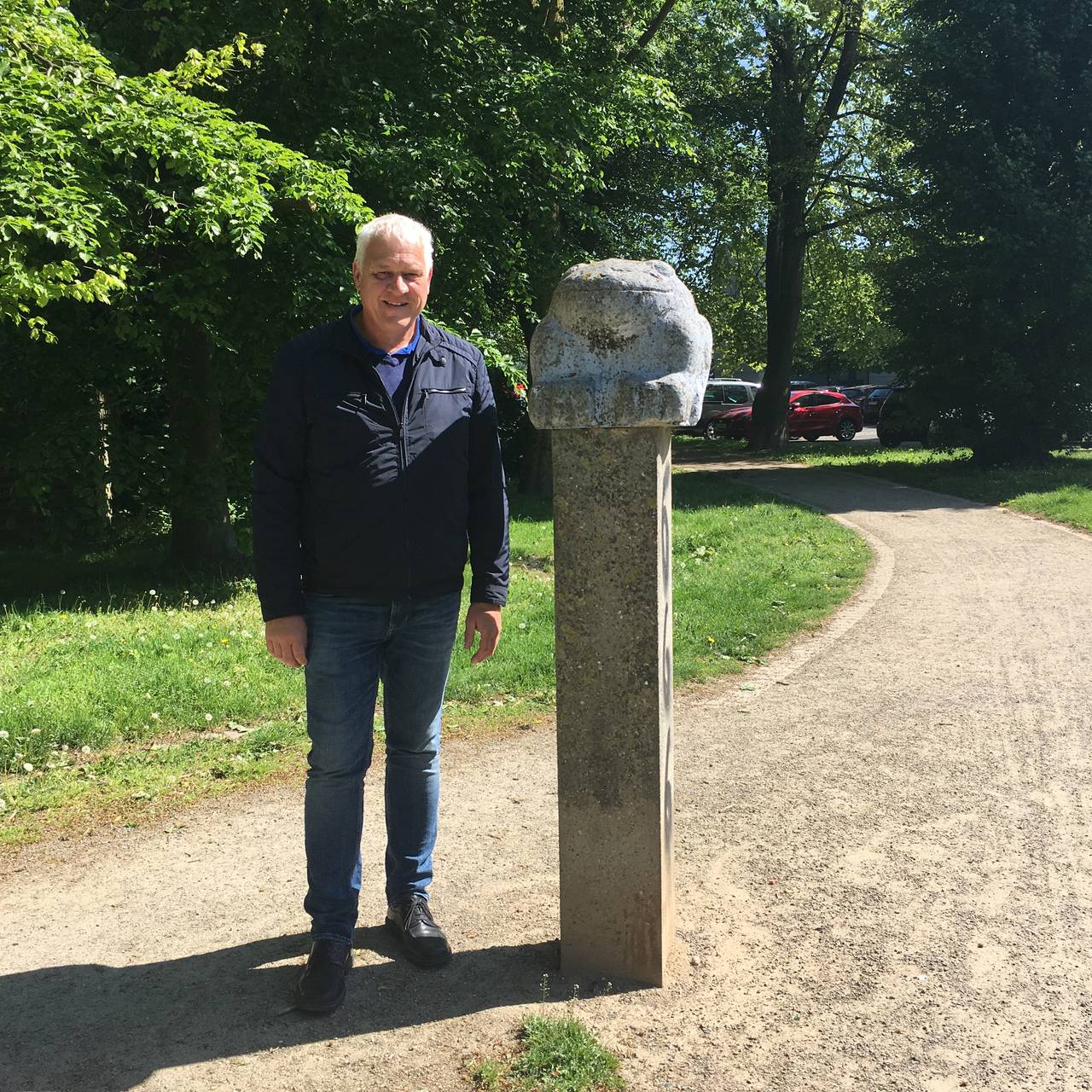 Ein älterer Mann, Landschaftsarchitekt Ulrich Falke, steht neben einem Steinfrosch auf einem Sockel in Schönebeck.