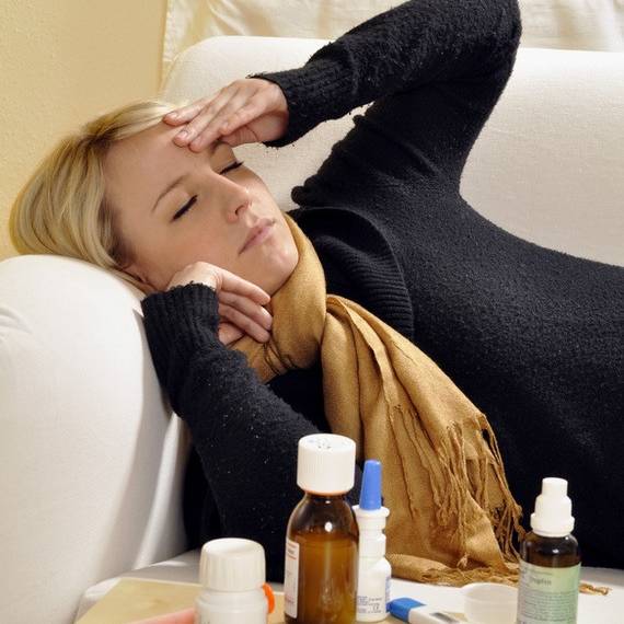 Eine blonde Frau liegt mit Schal auf dem Sofa und fasst sich an die Stirn. Vor ihr stehen ganz viele Medikamente.