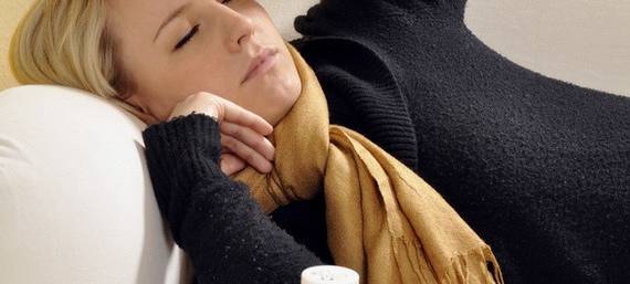 Eine blonde Frau liegt mit Schal auf dem Sofa und fasst sich an die Stirn. Vor ihr stehen ganz viele Medikamente.