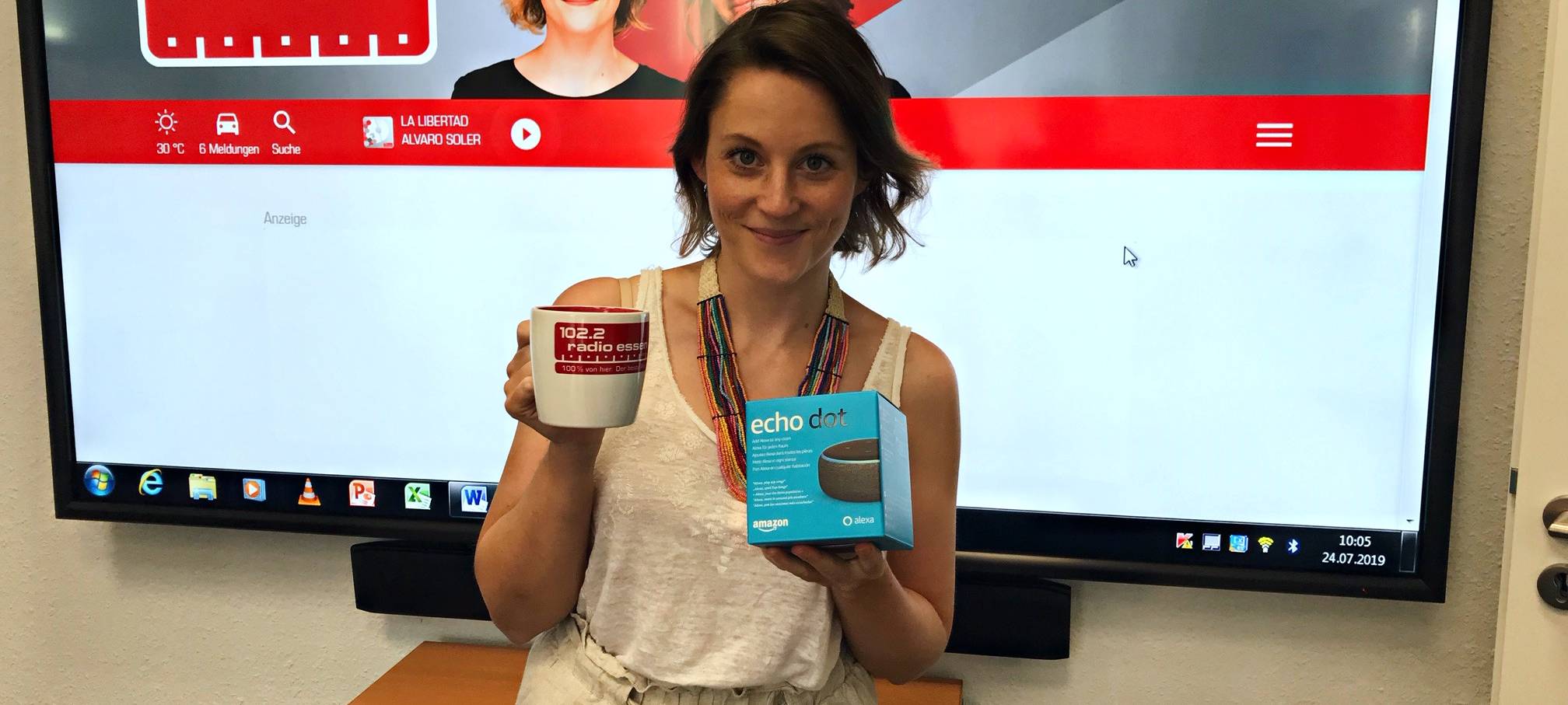 Radio Essen-Morgenmoderatorin Teresa Ledabyl steht lächelnd vor einem großen Fernseher, der die neue Radio Essen-Website zeigt. Sie hält eine weiß-rote Tasse von Radio Essen in der Hand und eine Alexa.