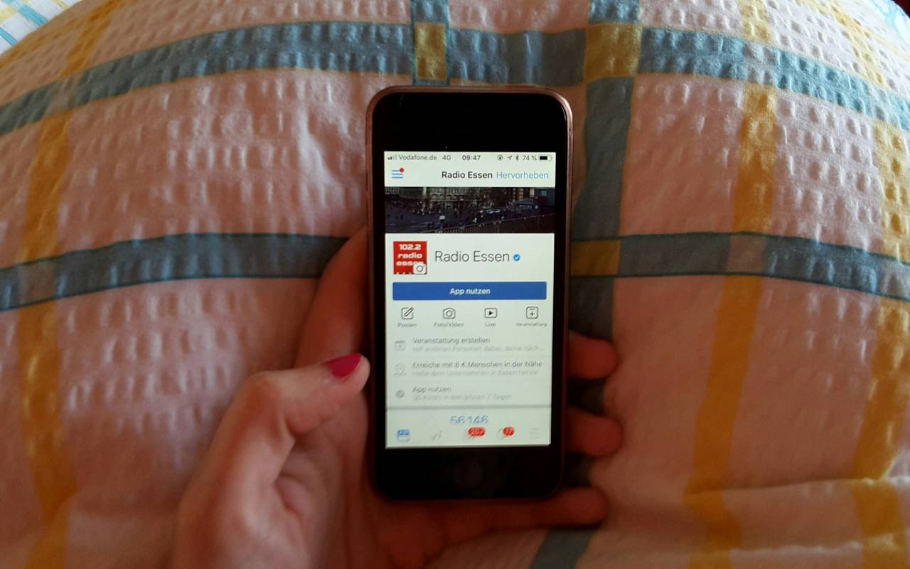 Eine Hand hält ein Smartphone mit der Radio Essen-Facebookseite über einer Bettdecke