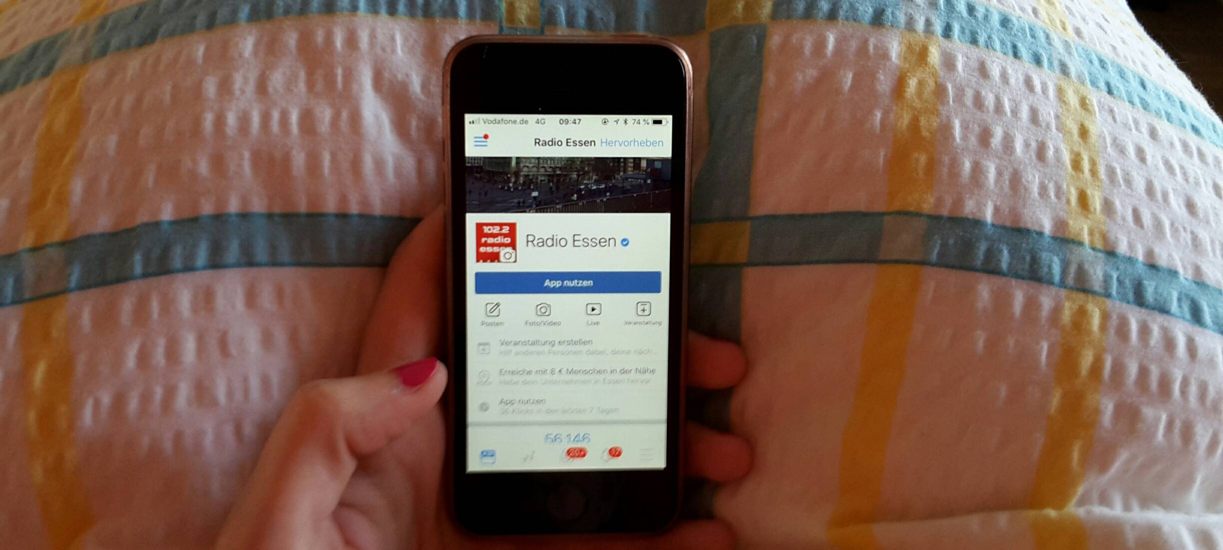 Eine Hand hält ein Smartphone mit der Radio Essen-Facebookseite über einer Bettdecke