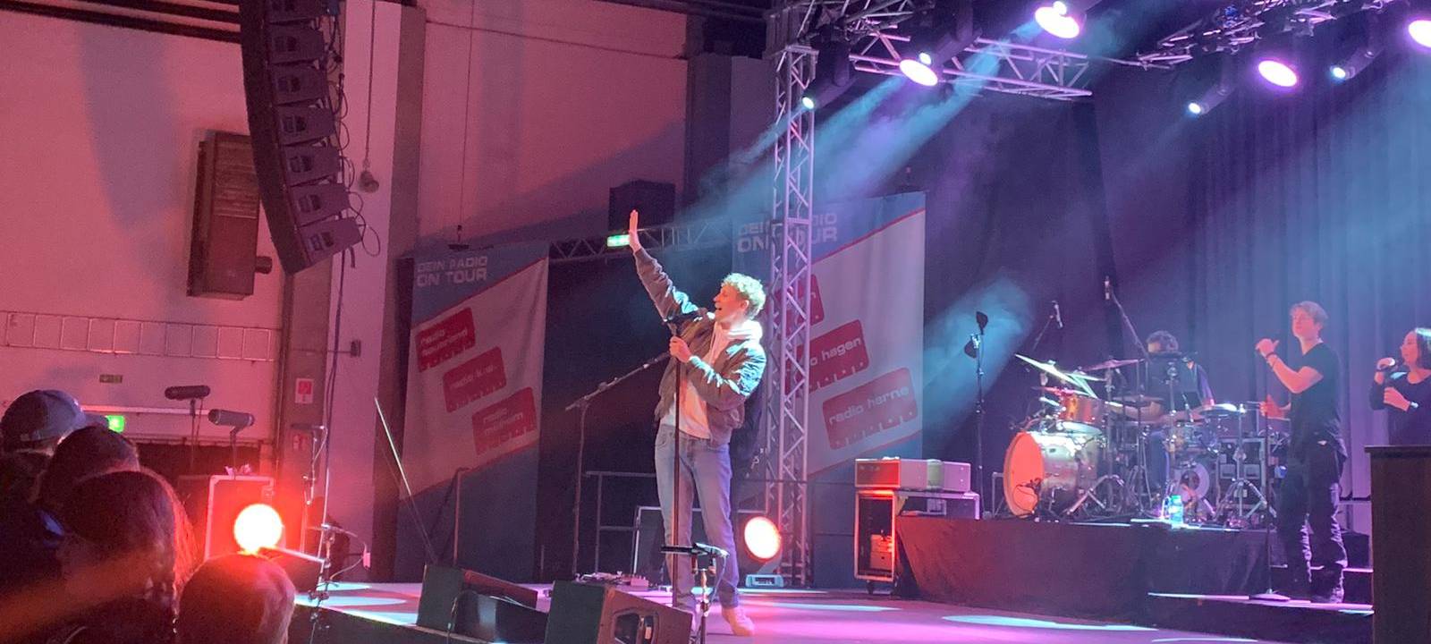 Blonder Mann steht mit hochgestrecktem Arm auf einer Bühne