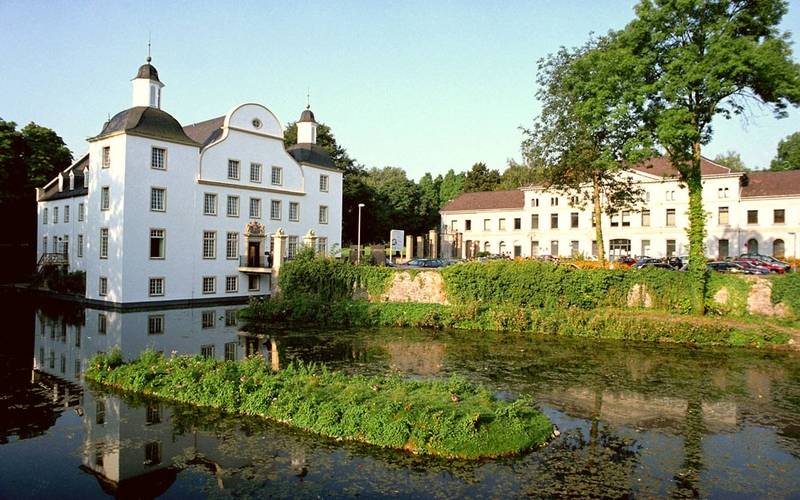Das Schloss Borbeck ist ein weißes großes Schloss mit hohen Fenstern, direkt an einem kleinen See gelegen mit einem Park dahinter.