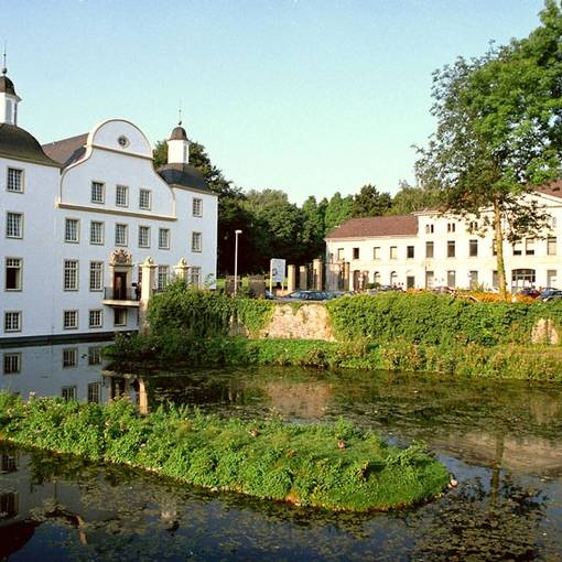 Das Schloss Borbeck ist ein weißes großes Schloss mit hohen Fenstern, direkt an einem kleinen See gelegen mit einem Park dahinter.